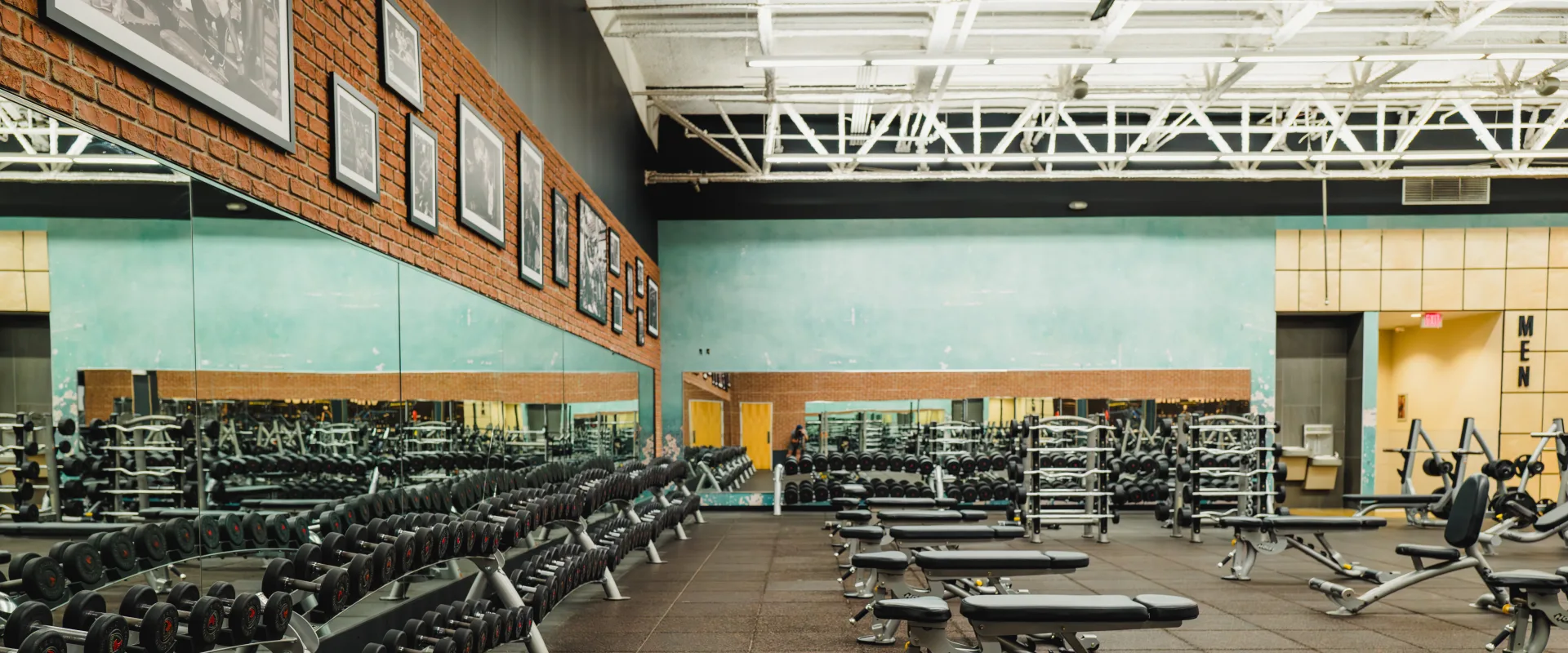 Gold's Gym dumbbell section with rows of neatly racked weights, benches, large mirrors, brick accents, and a teal wall in the background.