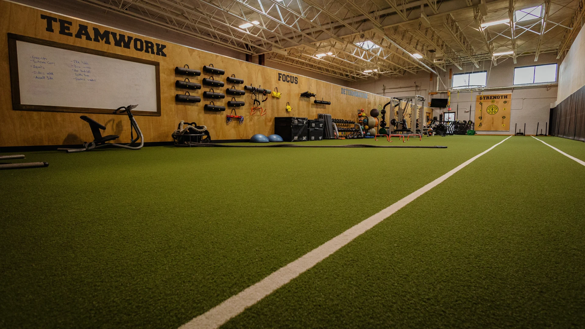 Turf area at Gold's Gym New York (Poughkeepsie), featuring a spacious green turf lined with white markings for agility drills and sled pushes. The wall displays motivational words like “TEAMWORK,” “FOCUS,” and “STRENGTH,” alongside functional training equipment including sandbags, battle ropes, medicine balls, and resistance bands. A whiteboard with workout notes adds a dynamic coaching element, while natural light filters through high windows, creating an energetic training environment.