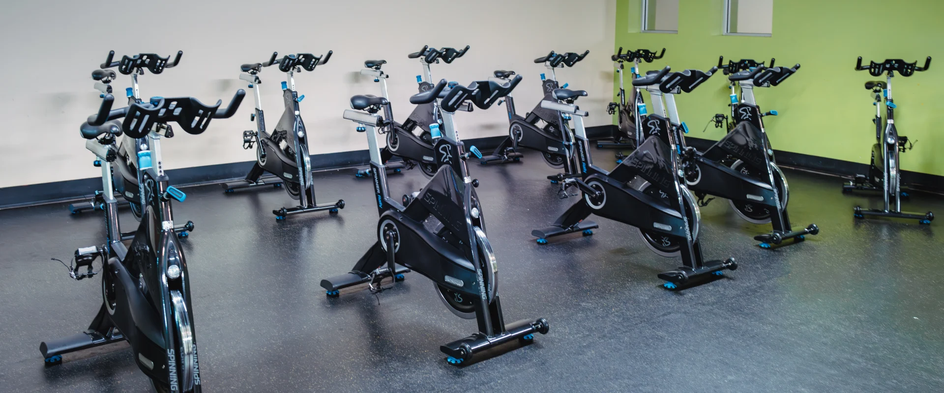 Group cycle studio at Gold's Gym Austin (Bee Caves) featuring rows of black stationary bikes arranged on a black floor. The room has light-colored walls with a green accent and a wall clock above. Two windows and a mounted fan are visible, adding ventilation and natural light to the space.