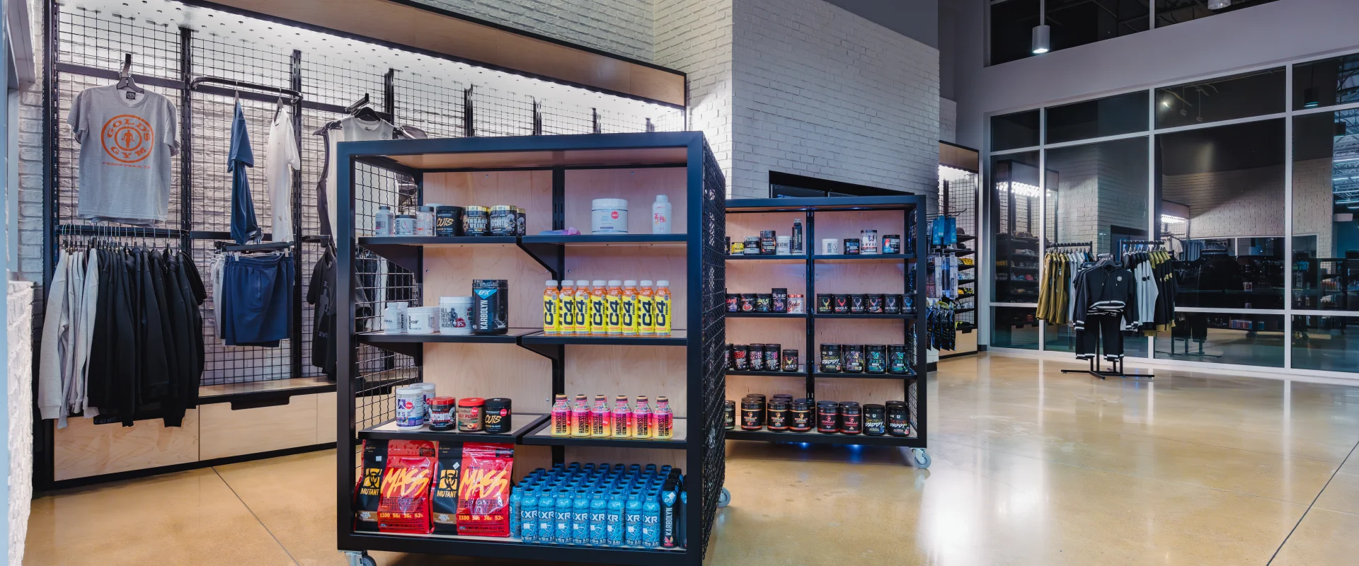 A Gold's Gym pro shop with shelves displaying supplements, energy drinks, and protein powders, alongside racks of branded apparel in a modern, open space.