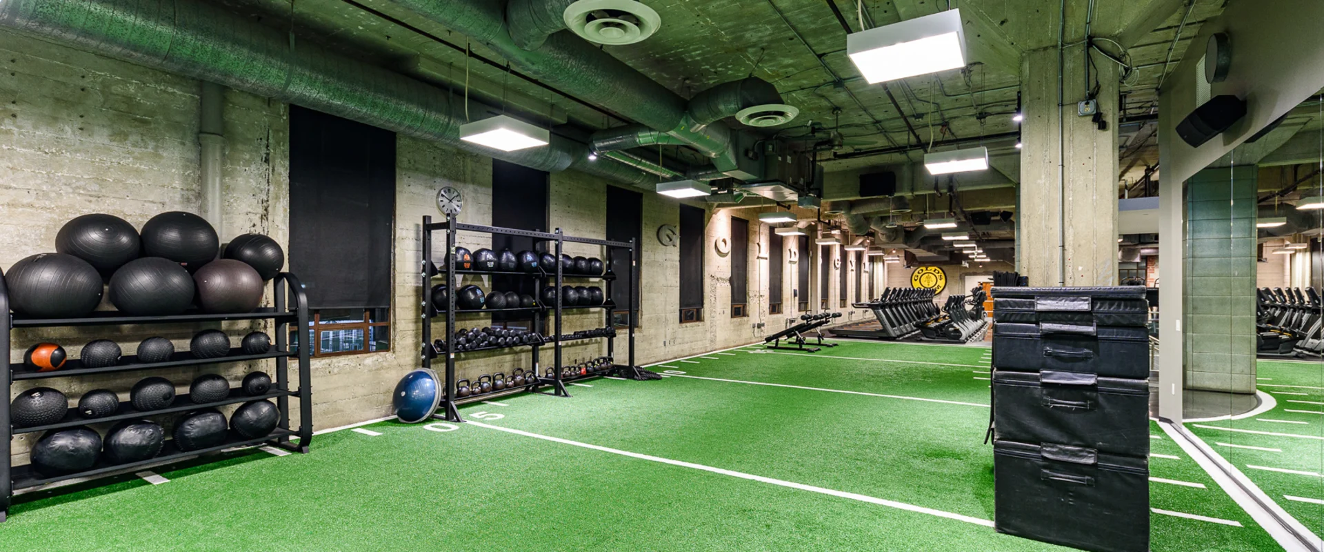 Turf area at Gold’s Gym Hollywood, featuring a green artificial turf space marked with white lines for functional training. Black medicine balls and kettlebells are neatly organized on racks along a concrete wall. A stack of black soft plyometric boxes is positioned near a mirrored wall. Exposed ductwork and industrial lighting fixtures line the ceiling. In the background, adjustable benches and strength training equipment are visible. A Gold’s Gym logo is displayed on a distant wall.