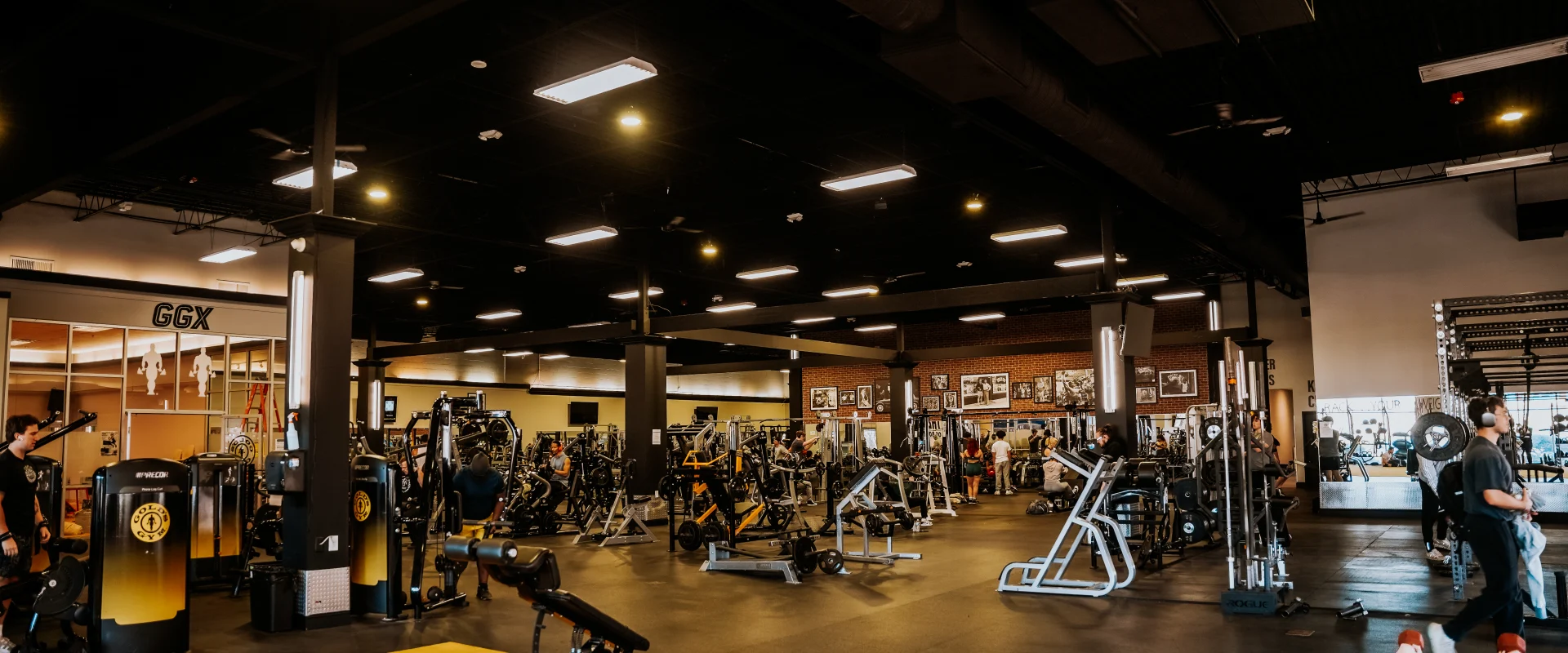 Machine weights area at Gold's Gym Murfreesboro, Tennessee, featuring rows of strength training equipment on a black floor, with a yellow carpet section in the foreground, high ceilings, and framed photos on a brick accent wall in the background.