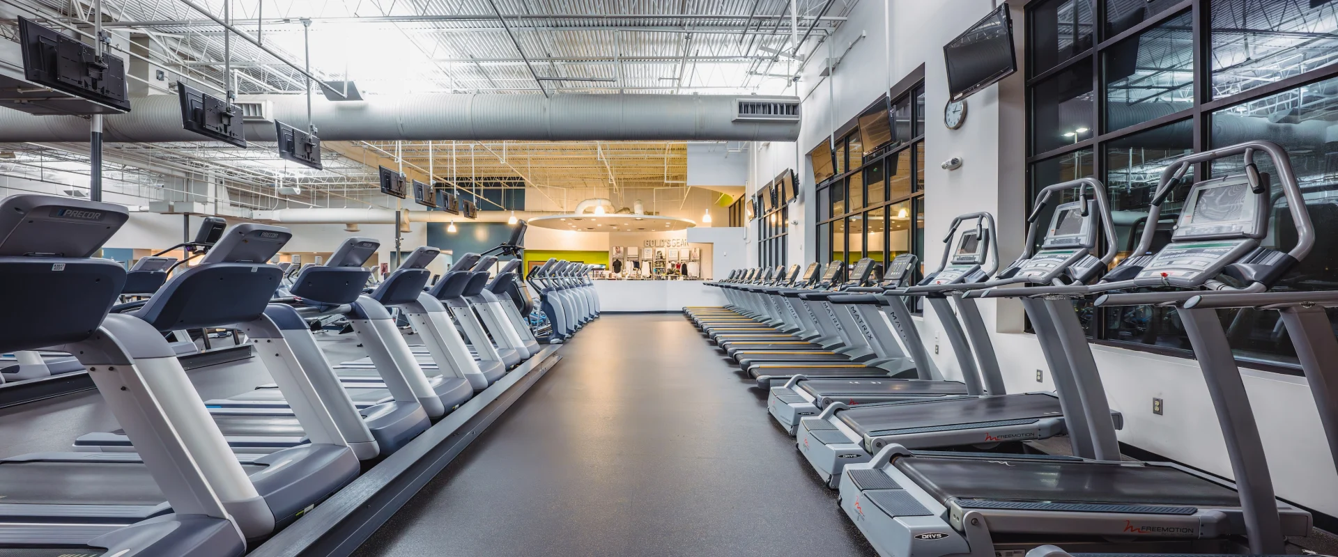 Spacious gym interior with rows of unoccupied treadmills, bright overhead lighting, large windows, and a modern, clean atmosphere.