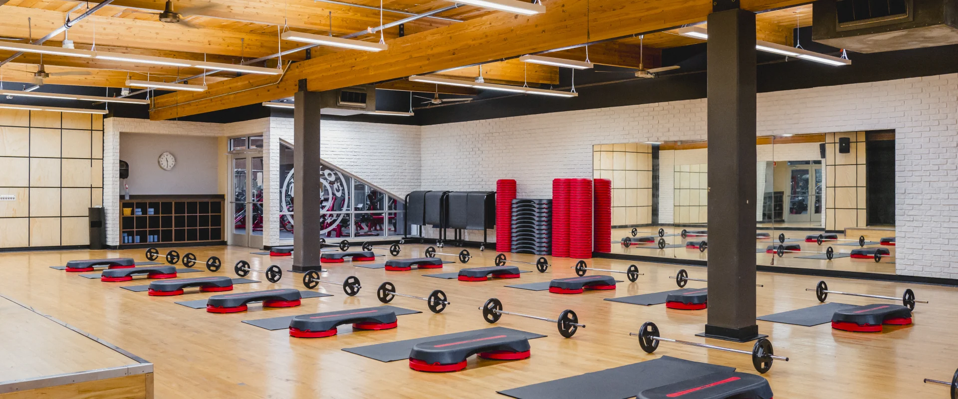 A bright group exercise studio with wooden floors, mirrored walls, a wooden ceiling, and neatly arranged steps, barbells, and mats for workout classes.