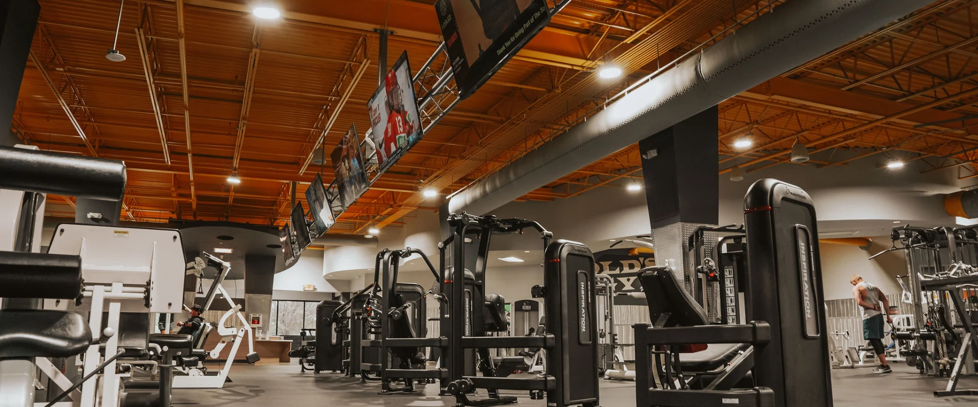 Machine weights area at Gold’s Gym, Chapman, Tennessee, featuring a variety of strength-training machines aligned in neat rows under a high, orange-lit industrial ceiling. Overhead monitors display sports content, adding energy to the space. The equipment, including seated chest presses, leg curl machines, and more, is designed for targeted muscle workouts. A member is seen actively using one machine, while another adjusts weights in the background. The gym’s open layout and well-lit environment enhance accessibility and visibility for users.