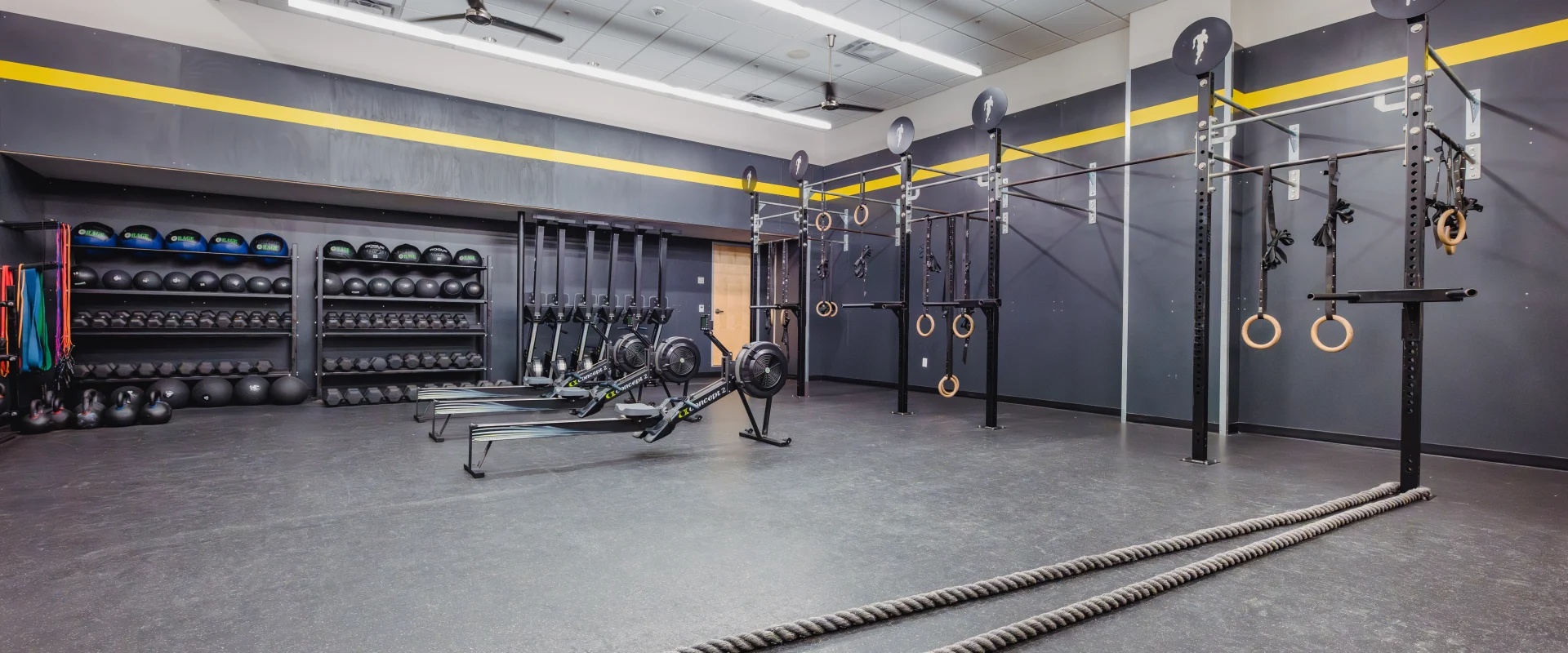Gold's Fit® room at Gold's Gym Austin (Downtown) featuring a functional training space with dark walls, yellow accents, and ceiling fans. The room includes pull-up rigs, gymnastic rings, battle ropes, rowing machines, and shelves stocked with kettlebells and medicine balls, creating a focused workout environment.