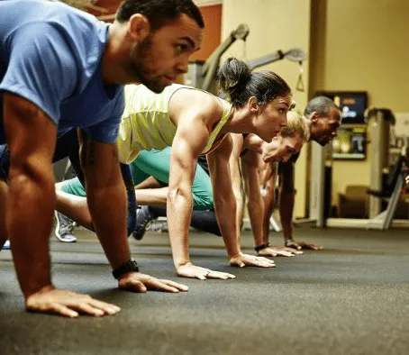 A group of people in athletic clothing are performing a plank exercise together in a fitness center, with various equipment and machines visible in the background.