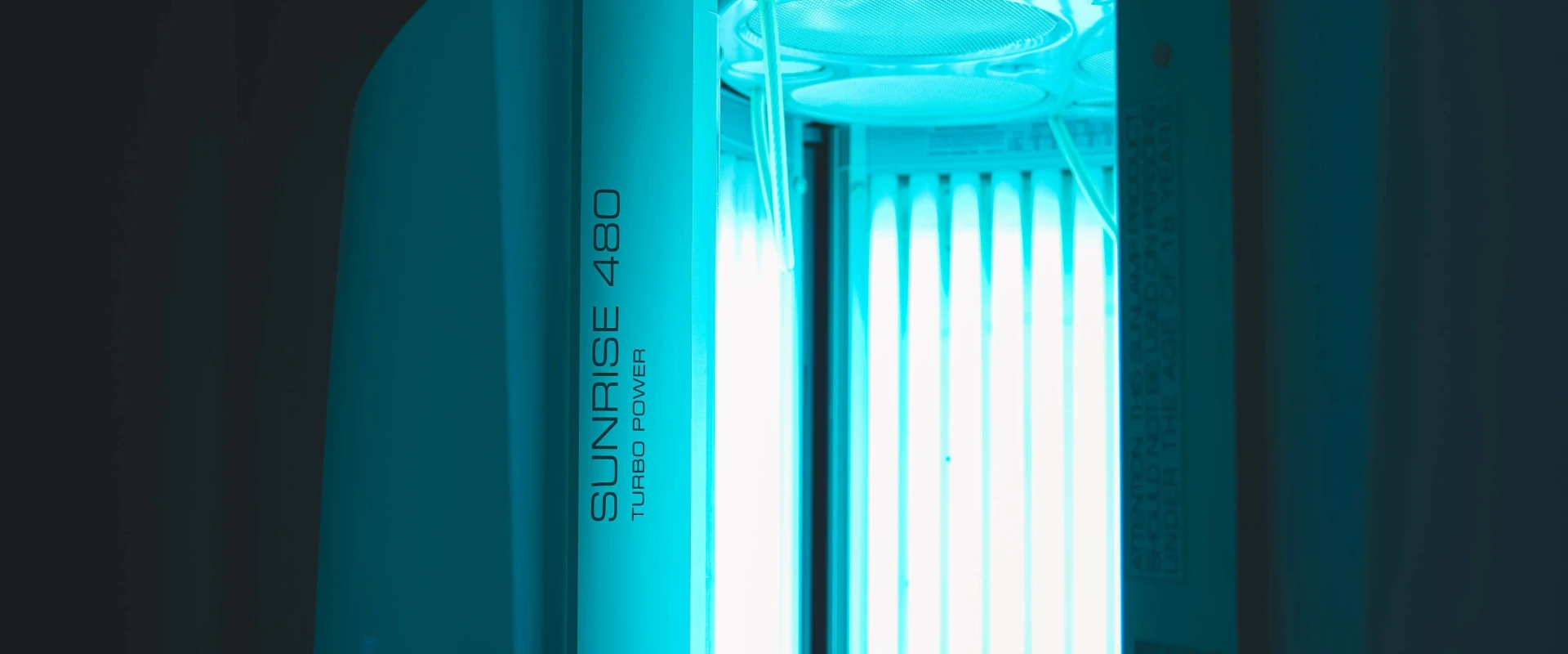 Gold's Gym Memphis, Tennessee (White Station) tanning area featuring an illuminated vertical tanning booth labeled "Sunrise 480 Turbo Power." The tanning bed emits a bright blue glow from its vertical UV light tubes. The interior fan and ventilation system are visible at the top. The partially open door reveals the sleek design and standing layout of the unit. The surrounding area is dimly lit, contrasting with the bright UV lights inside the booth.