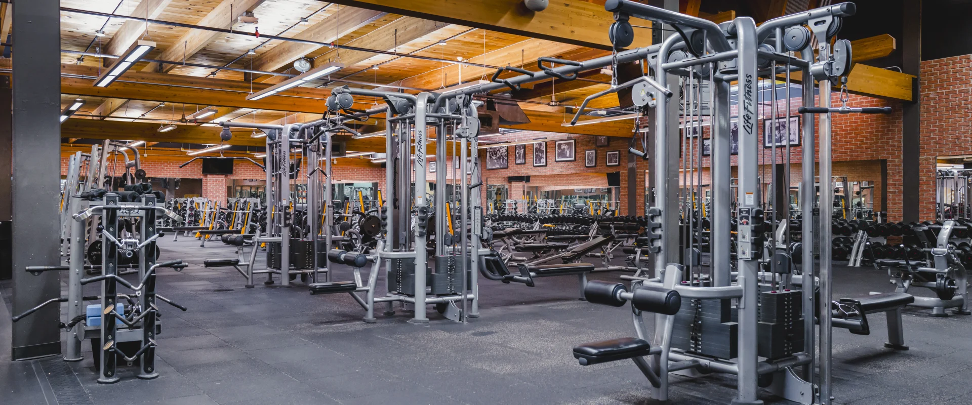 A gym area featuring cable machines and weightlifting equipment, surrounded by mirrored walls, exposed brick, and a wooden ceiling with beams.