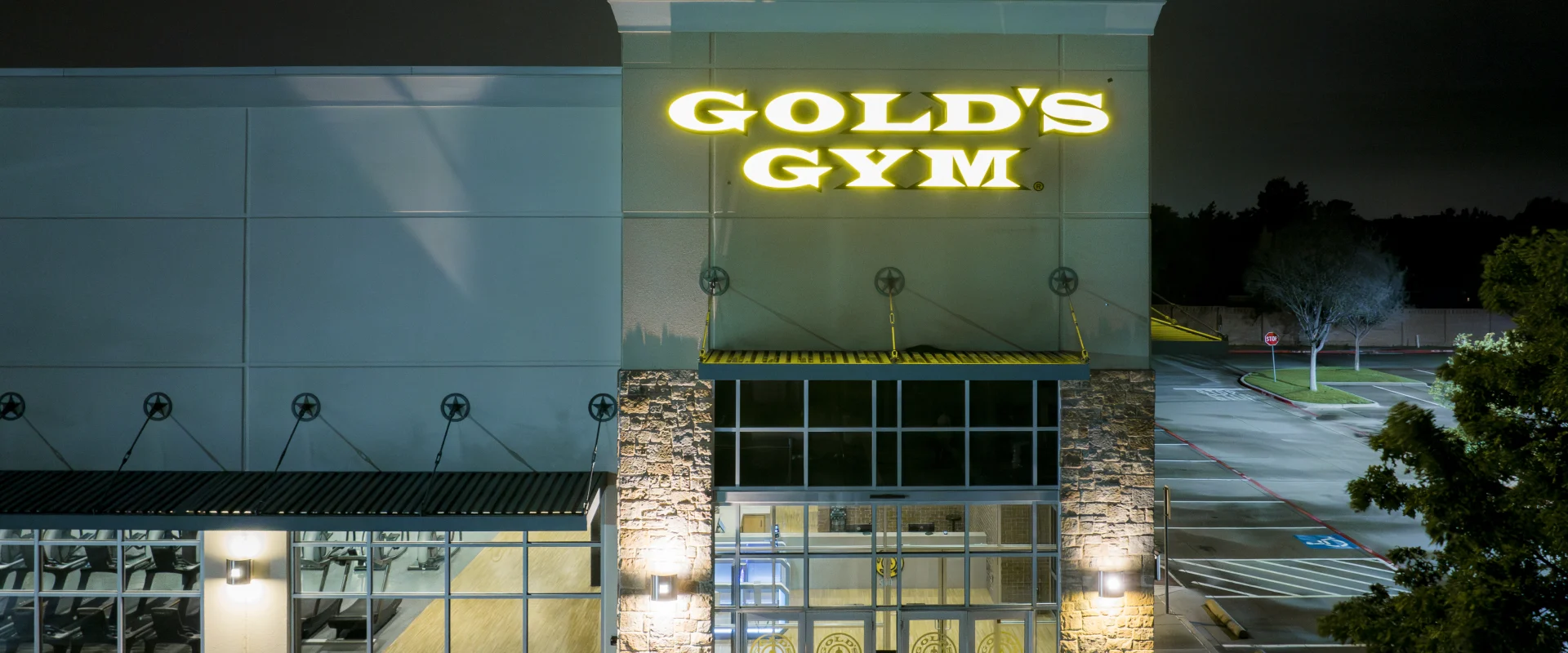 Exterior of Gold's Gym Little Elm, featuring a well-lit facade with a glowing yellow Gold's Gym logo. The entrance showcases glass doors with the gym's emblem, framed by stone pillars and large windows revealing workout equipment inside. The surrounding area includes a clean sidewalk and parking lot under the soft glow of exterior lighting.