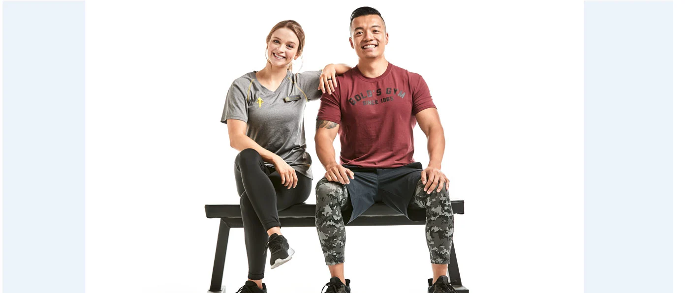 Two people, a man and a woman, are sitting on a bench against a plain white background, both smiling and appearing relaxed.