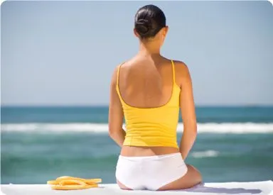 A person in a yellow tank top and white shorts sits on a beach, facing the ocean with waves crashing in the background.