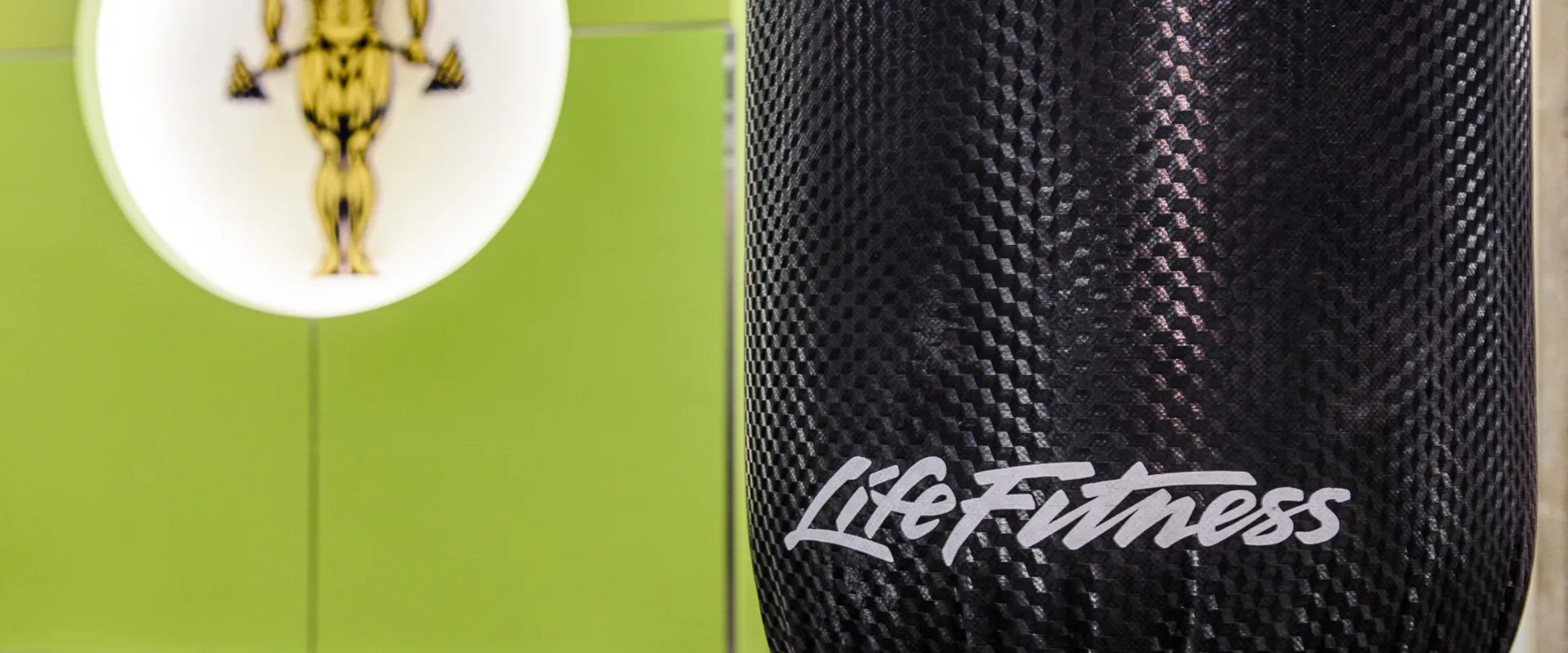 Boxing area at Gold’s Gym Long Beach, featuring a Life Fitness heavy bag with a textured black surface. The bag hangs in front of a green accent wall, where the gym’s iconic logo glows in the background.