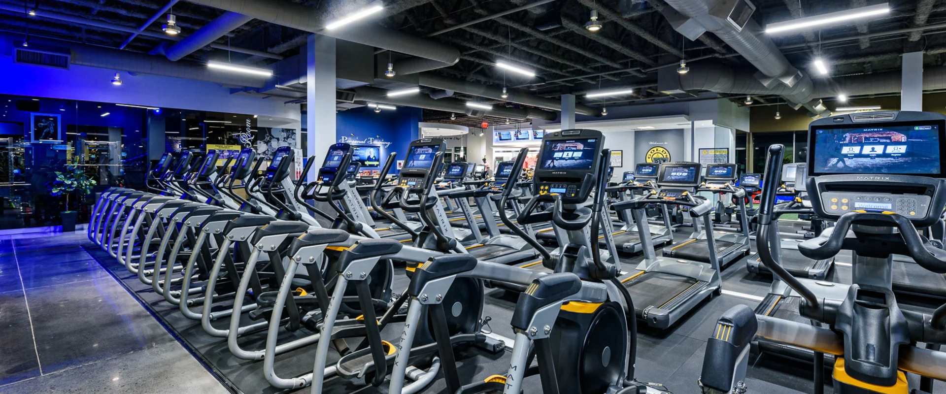 Cardio area at Gold’s Gym Thousand Oaks, featuring rows of elliptical machines and treadmills arranged on black rubber flooring. Each machine has a digital screen. Overhead LED lighting illuminates the space, with an exposed industrial-style ceiling featuring ductwork. A Gold’s Gym logo is visible in the background, along with additional workout areas and a personal training section. Blue accent lighting highlights a nearby glass-walled area.
