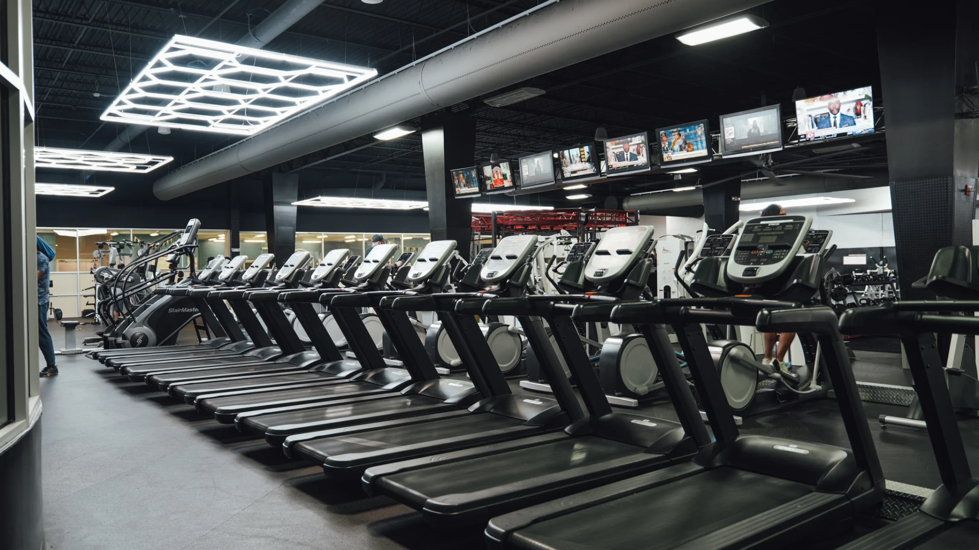 A row of high-end treadmills and elliptical machines at Gold's Gym Brassfield, positioned under sleek industrial lighting. Multiple overhead TVs display news and sports channels, creating an engaging workout environment. The modern black-and-gray gym aesthetic is complemented by exposed ductwork and geometric LED ceiling lights.