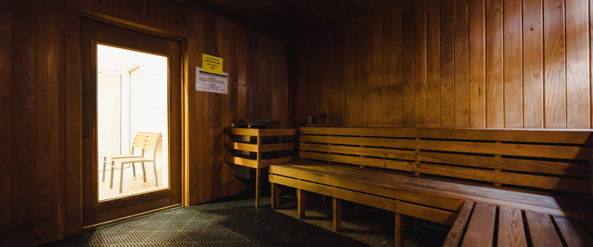 A wooden sauna room at Gold's Gym with built-in bench seating, warm lighting, and a door leading to a well-lit adjacent area with additional seating.
