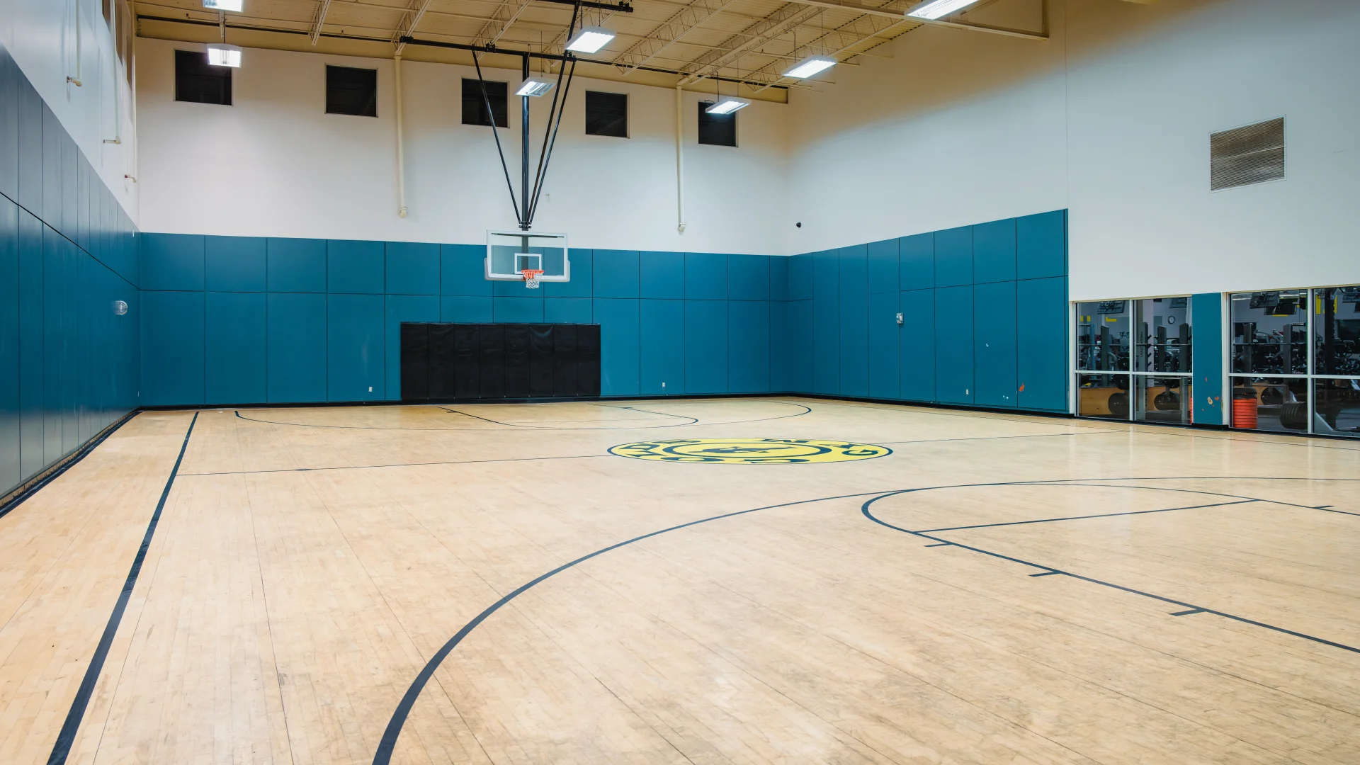 A basketball court at Gold's Gym in Austin (Hesters Crossing) featuring a polished wooden floor with court markings, including the gym's logo at the center. The walls are padded in blue with a mounted basketball hoop on one end, and high ceilings with bright overhead lights illuminate the space. A glass wall on the right side provides a view into the adjacent fitness area.
