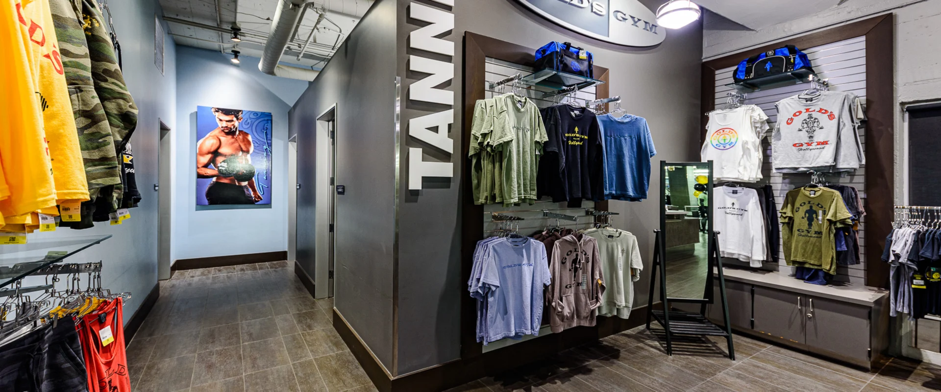 Pro shop at Gold’s Gym Hollywood, featuring a variety of gym apparel displayed on racks and shelves. T-shirts, hoodies, and tank tops with Gold’s Gym branding are arranged alongside gym bags. A freestanding mirror is positioned nearby. The space includes a dark gray wall with bold "TANNING" lettering, leading to a hallway with tanning rooms. A framed Gold’s Gym logo is mounted above the apparel section. In the background, a large poster of a bodybuilder is visible. Overhead lighting and an exposed ceiling highlight the space.