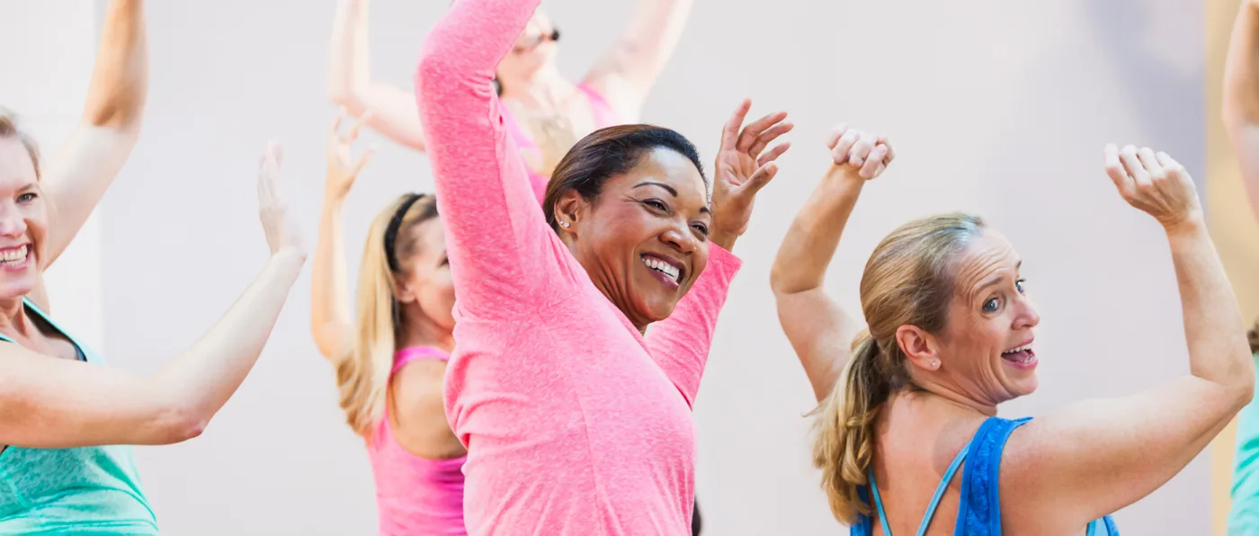 Three women in colorful workout clothes are enthusiastically raising their arms in a fitness or dance class setting.