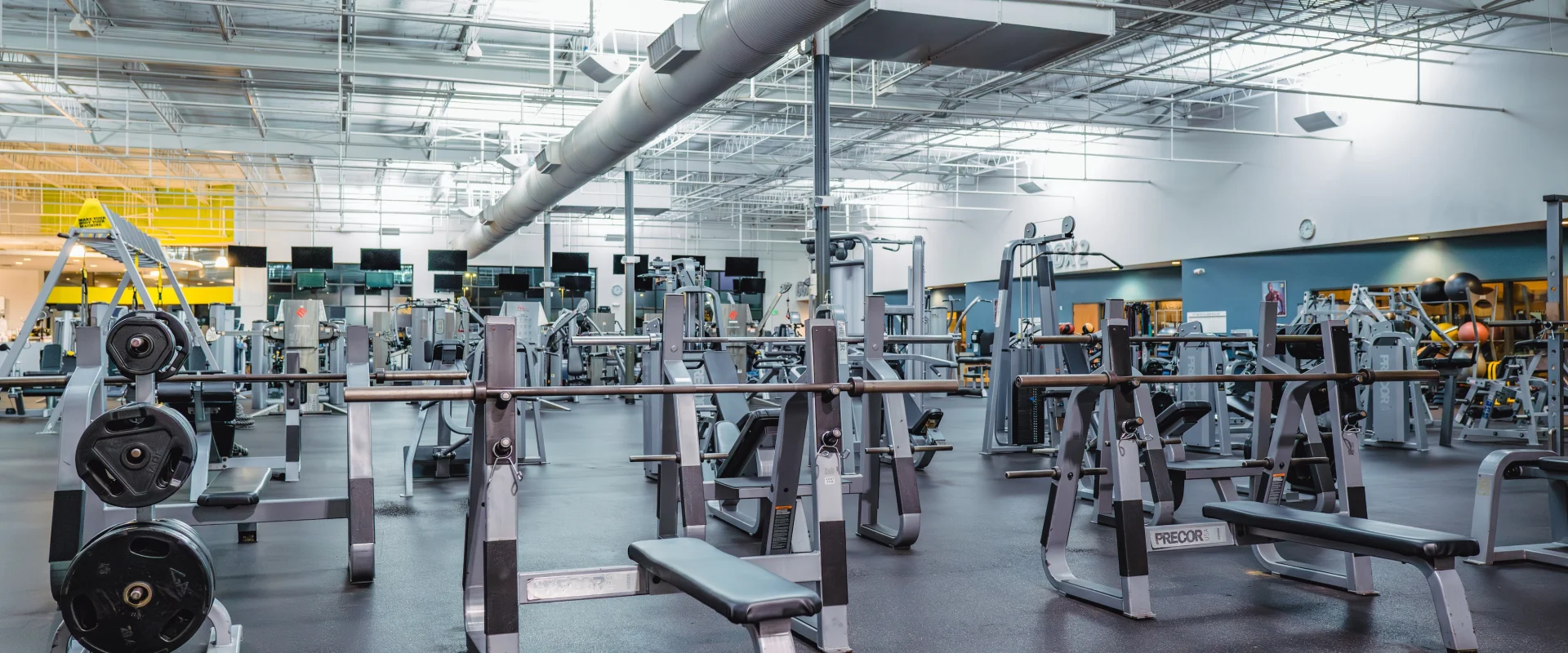 Spacious gym with various weightlifting equipment, including benches and barbells, under bright lighting, conveying a clean and organized atmosphere.