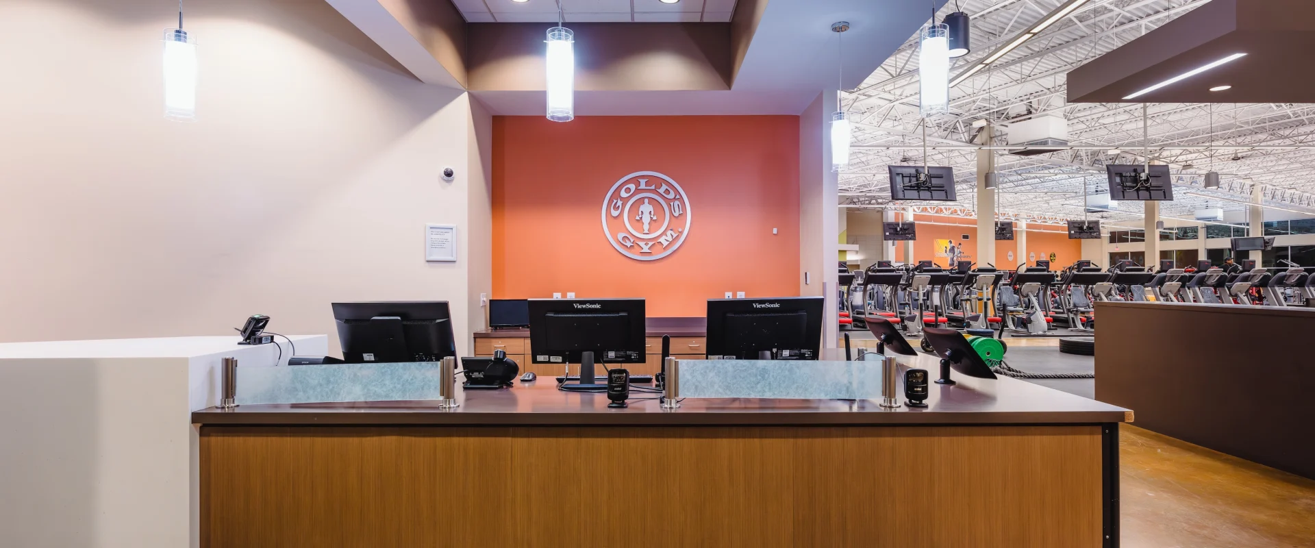 Reception desk of a gym with warm lighting and modern decor. Behind the desk, a wall features a gym logo. Exercise machines are visible in the background.