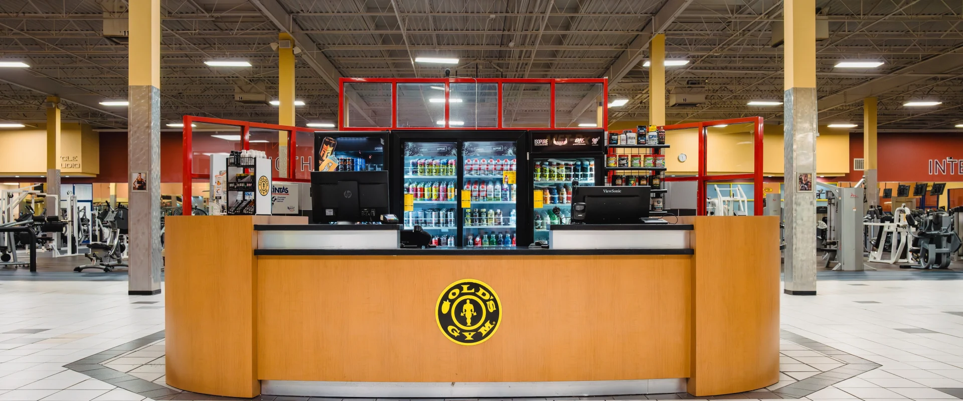 Spacious gym interior with a central reception desk. Behind it are shelves stocked with drinks and snacks. Weightlifting equipment is visible in the background.