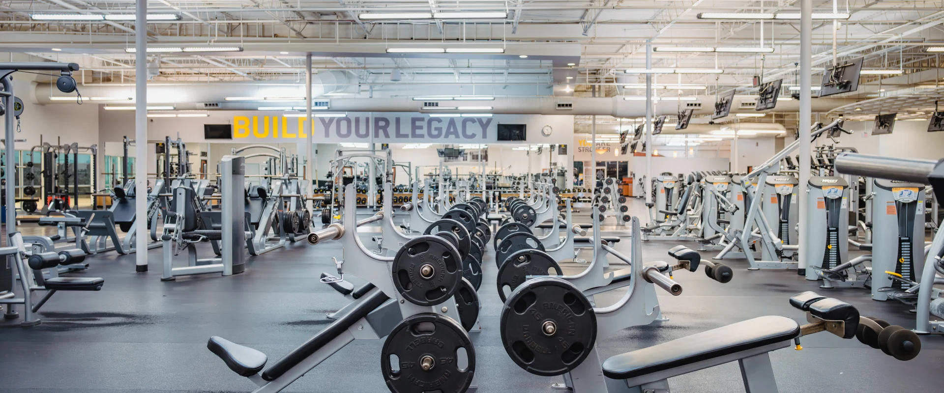 Free weight area at Gold's Gym in Austin (Hesters Crossing), featuring rows of benches with barbells and weight plates on black rubber flooring. The gym includes additional weightlifting equipment, mirrored walls, and high ceilings with exposed ducts. Motivational text "BUILD YOUR LEGACY" is prominently displayed on the wall, complementing the open, brightly lit layout filled with strength-training machines and free weights.