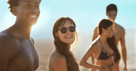 A smiling woman in sunglasses stands in the foreground, with two shirtless individuals visible in the background against a bright, sunny sky.