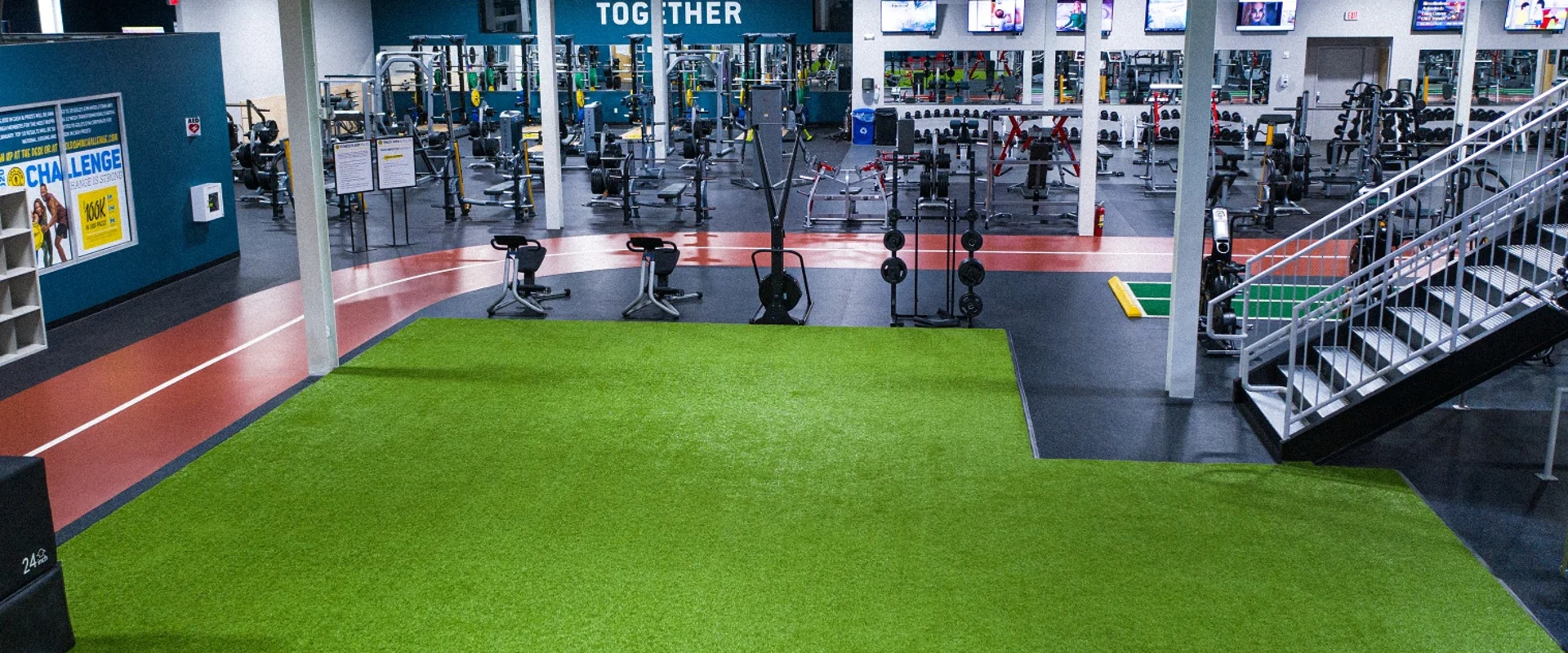 A large indoor gym with a green artificial turf floor, various exercise equipment, and a "Together" sign on the wall in the background.