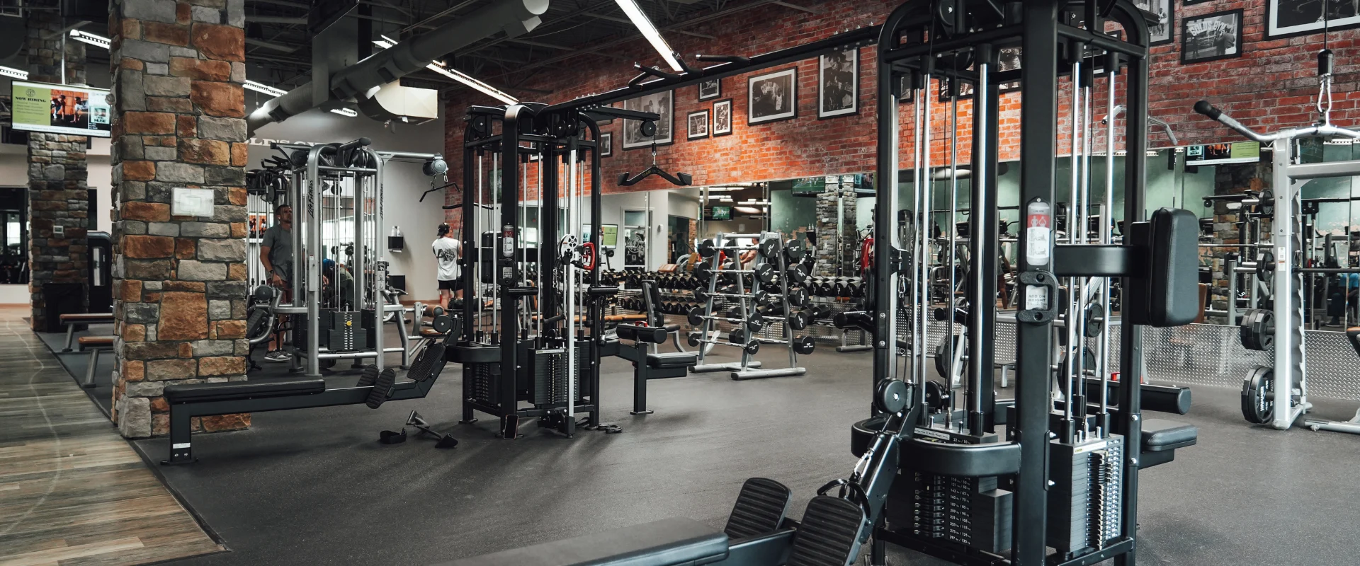 The free-weight section at Gold's Gym Rock Hill features black dumbbells neatly arranged on multi-tiered racks, surrounded by adjustable weight benches. A long mirrored wall reflects the space, while red brick accent walls display framed photos of legendary bodybuilders.