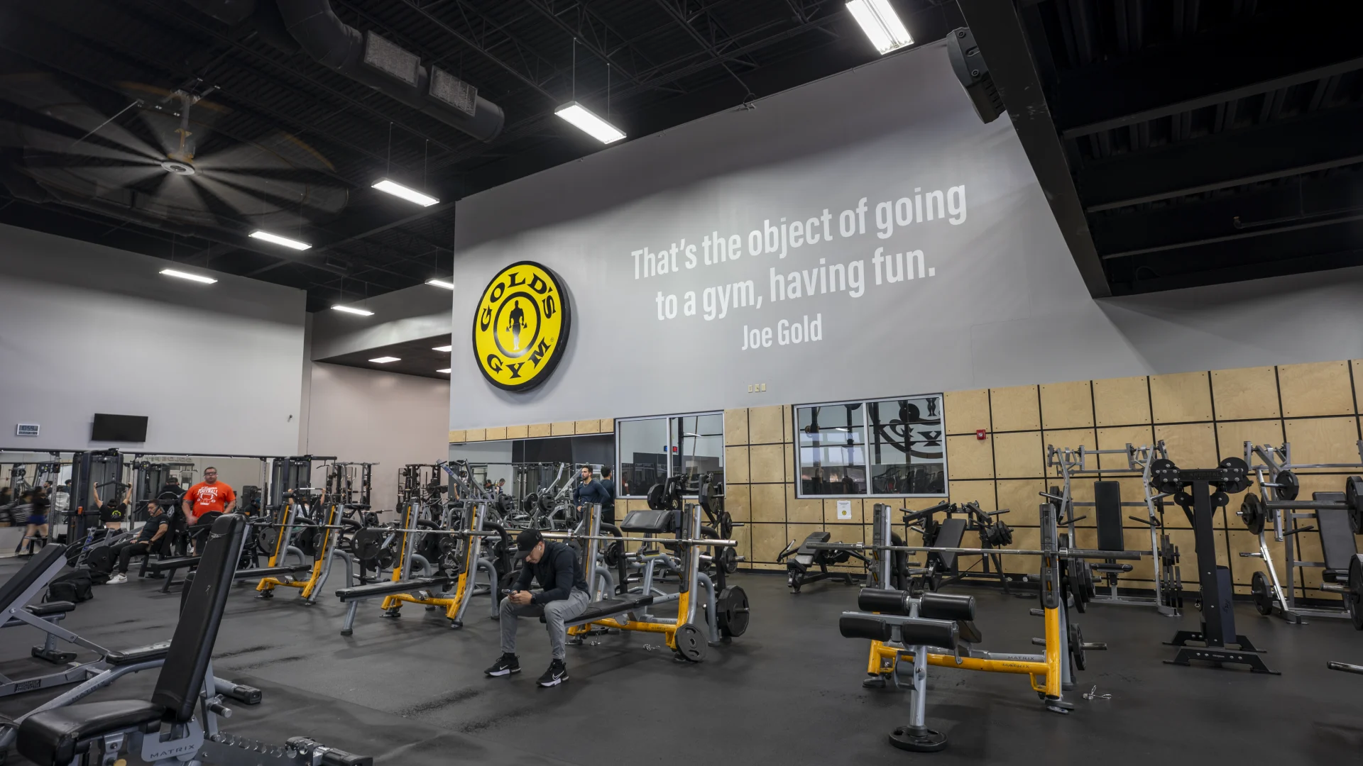 An interior gym wall featuring a large circular yellow and black Gold’s Gym logo on the left. To the right, a quote in white sans-serif lettering reads: "That’s the object of going to a gym, having fun. Joe Gold." Below the quote are interior windows and tan wood-paneled walls.