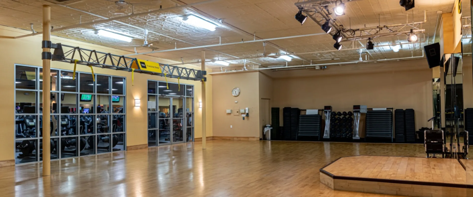Group exercise studio at Gold’s Gym in East Northport, New York, featuring a spacious hardwood floor, mirrored walls, and a raised instructor stage. The room includes stacked step platforms, weights, and mats along the back wall. Overhead lighting and a ceiling-mounted TRX suspension system provide a versatile space for various fitness classes, with large windows offering a view of the main gym area.