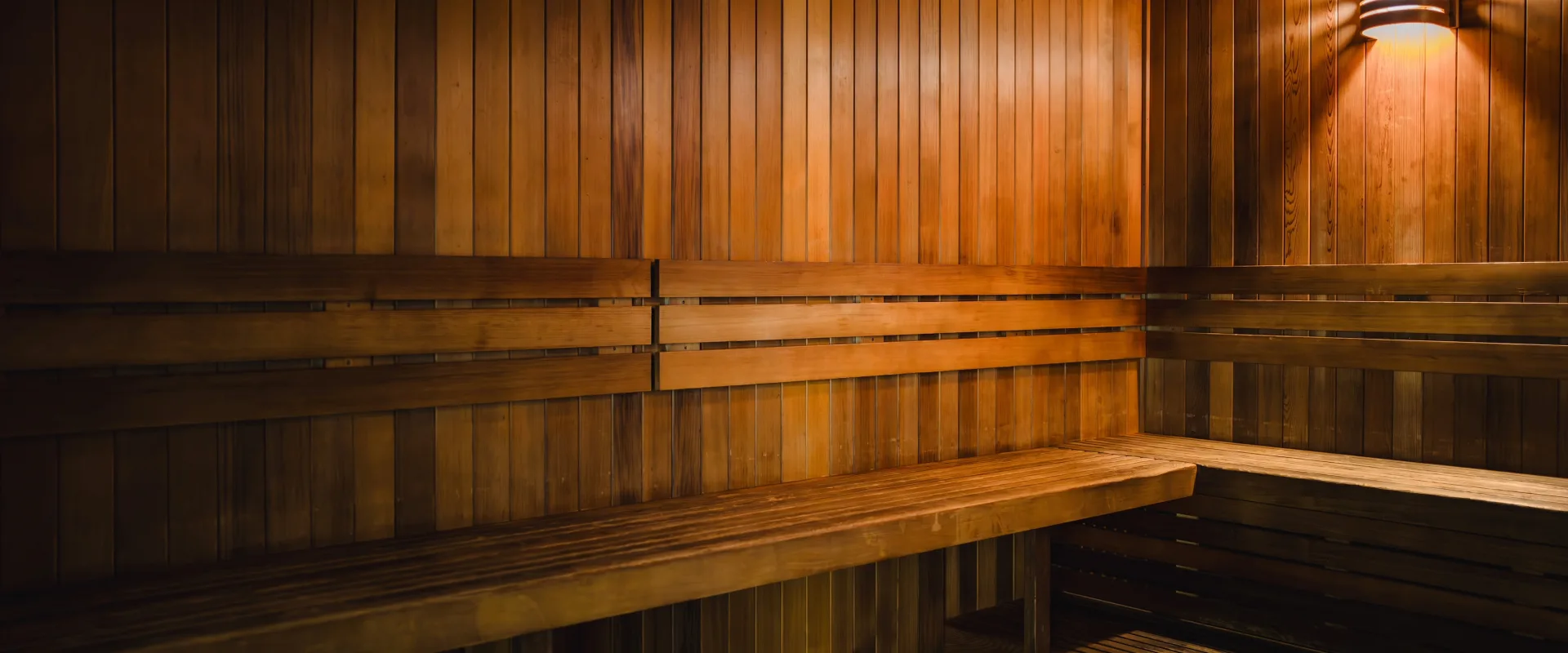 Sauna at Gold’s Gym in Texas (N. Round Rock), featuring wooden panel walls, tiered wooden benches, and a wall-mounted light fixture that casts a warm glow. The space includes upper and lower seating levels, providing options for different heat intensities. The clean, minimalist design emphasizes natural wood tones, creating a relaxing environment for post-workout recovery.