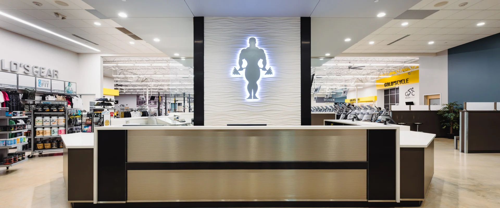 Front desk at Gold’s Gym in Texas (N. Round Rock), featuring a rectangular design with a brushed metal finish and black accents. A large illuminated silhouette of the Gold’s Gym logo is centered on a textured white backdrop behind the desk. To the left, the pro shop displays supplements, apparel, and accessories. The gym floor with cardio equipment and Gold’s Cycle area is visible in the background, with overhead lighting and polished concrete floors.