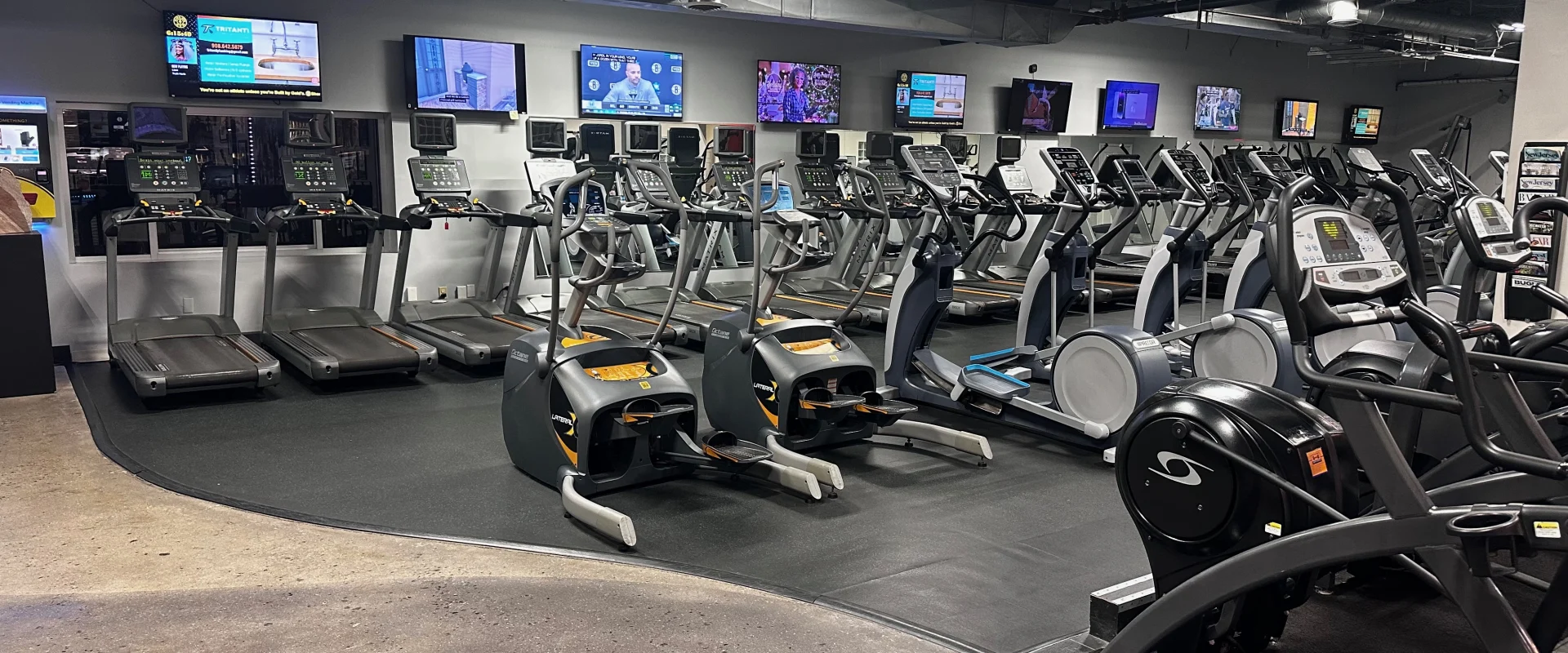 Gold's Gym Bridgewater cardio area featuring treadmills, elliptical machines, and stationary bikes lined up against a mirrored wall with multiple mounted televisions overhead. The dark ceiling contrasts with bright overhead lighting, and the rubber flooring defines the workout space.