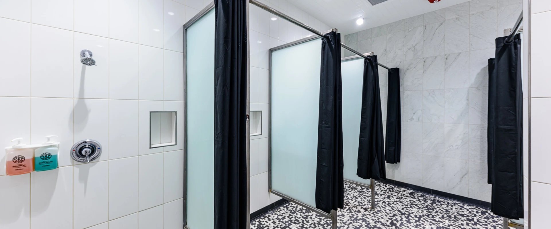 Gym shower area at Gold's Gym Garden Grove, featuring individual shower stalls with frosted glass panels and black curtains for privacy. The space has sleek white tiled walls with built-in product dispensers and marble-style accents. The floor is covered in small black-and-white mosaic tiles, and recessed ceiling lights provide bright illumination.
