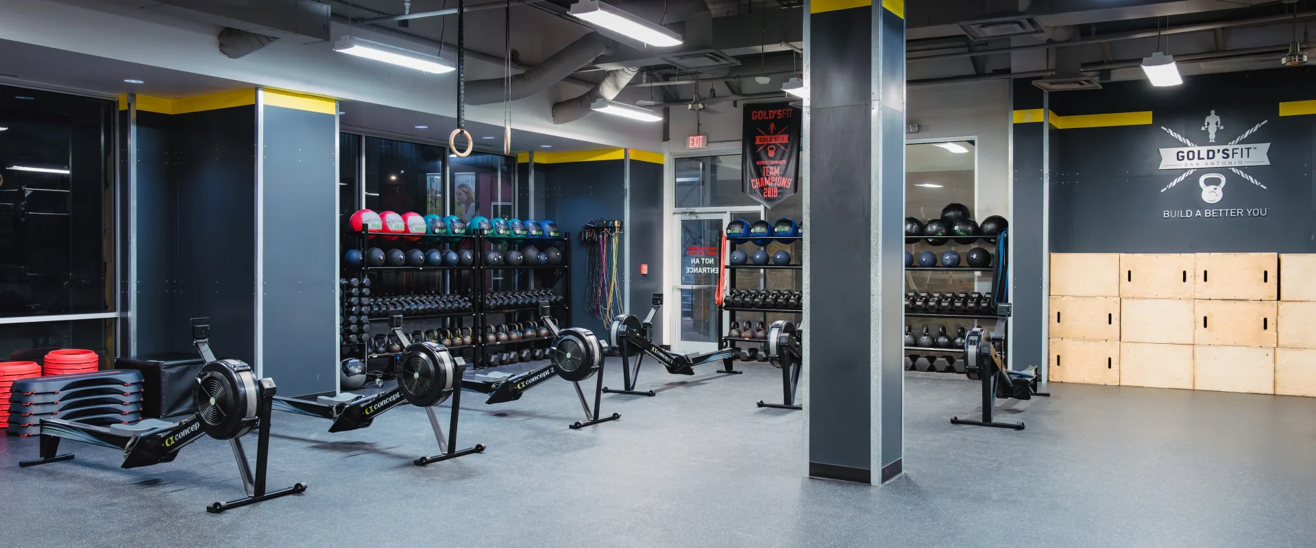 A modern gym with rowing machines, weight racks, medicine balls, and workout boxes. Bright lighting and sleek design create an energetic atmosphere.