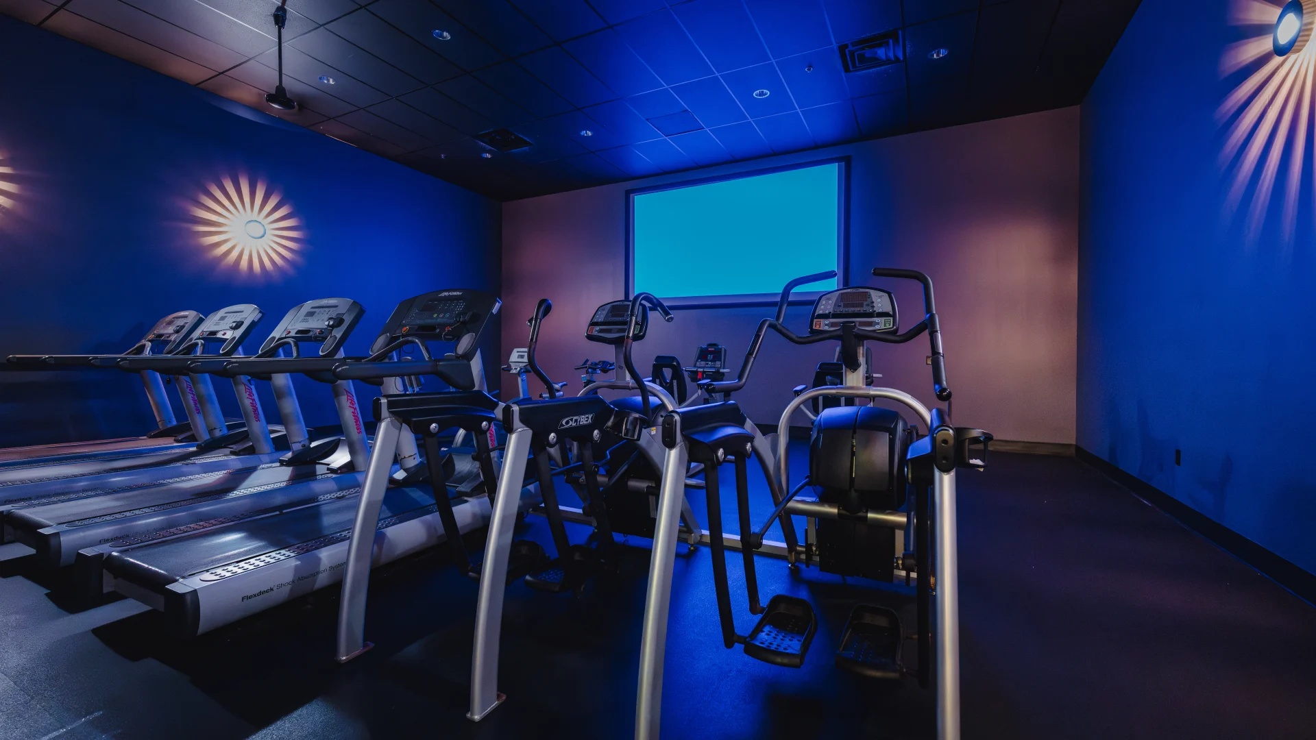 Cardio Cinema room at Gold’s Gym Austin (Anderson Arbor) featuring treadmills and elliptical machines facing a large projection screen. The room is dimly lit with wall-mounted lights creating a cinematic ambiance.