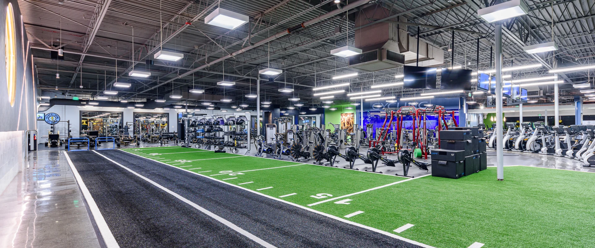Indoor functional training area at Gold's Gym Garden Grove, featuring a long sprint track with black and green turf marked with white yard lines. The space includes weighted sleds, plyometric boxes, battle ropes, and rowing machines. In the background, weightlifting stations and cardio machines line the gym floor, illuminated by bright overhead lighting.