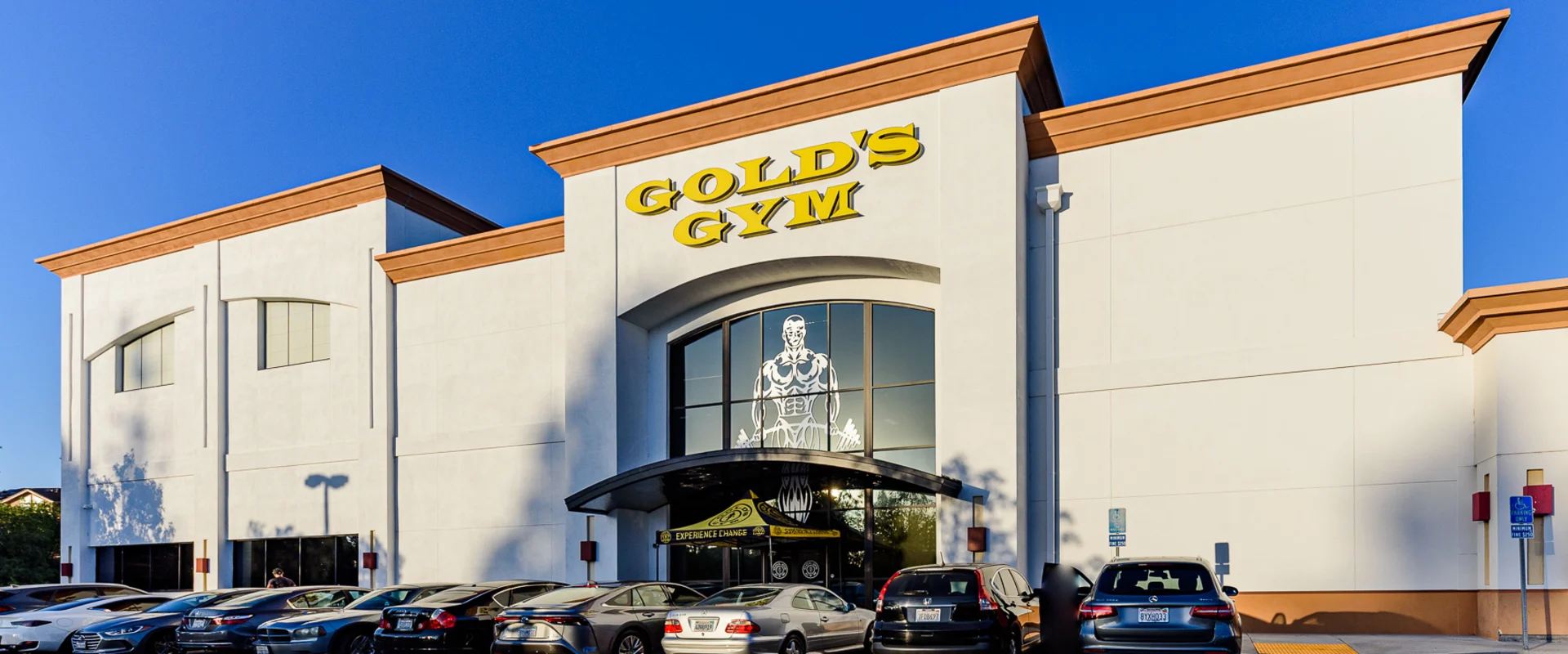 The entrance to Gold’s Gym La Mirada features a grand white facade with large yellow signage above the glass doors. A row of parked cars lines the front, with a bright blue sky in the background.