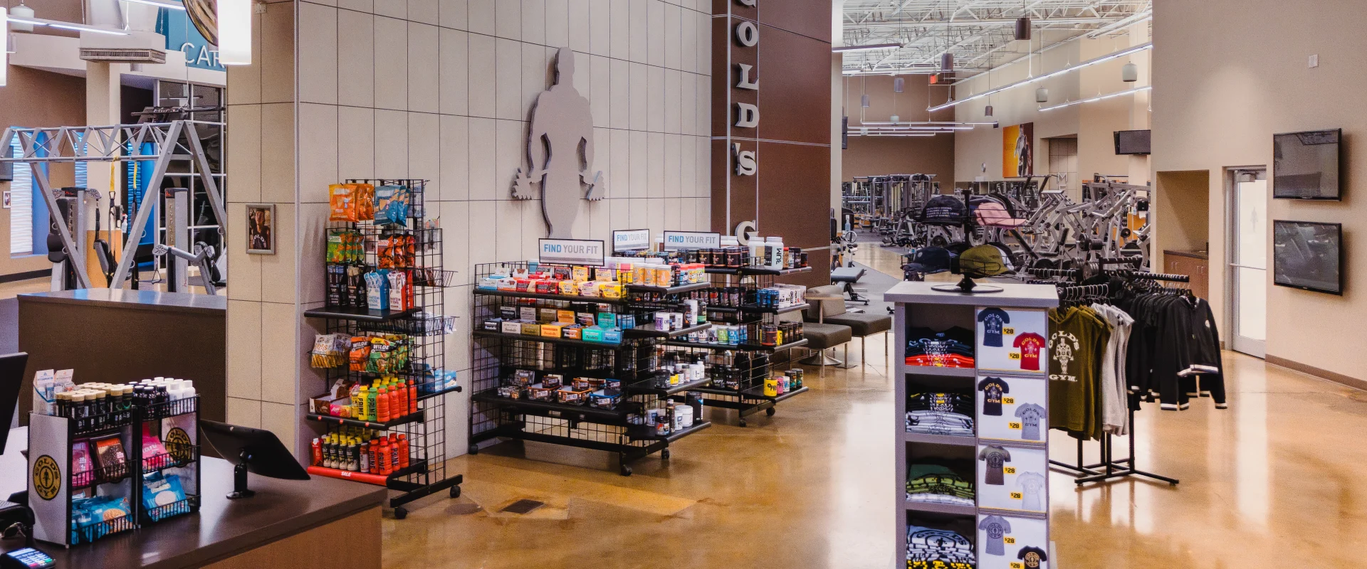 Pro shop at Gold's Gym Austin (Anderson Arbor) featuring racks with workout snacks, drinks, and supplements on the left, a display of gym apparel and accessories on the right, and workout equipment visible in the background under high ceilings.