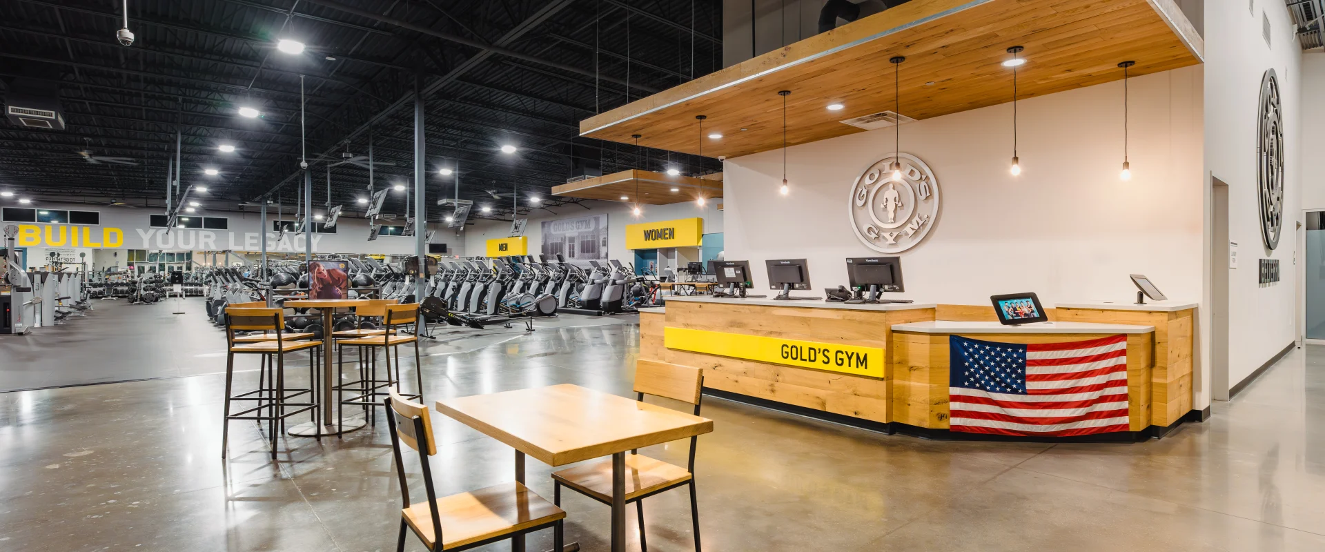 Gold's Gym front desk area with a wooden design featuring the gym's logo and an American flag. Nearby, a seating area with tables and chairs complements the open view of the gym floor filled with cardio and strength equipment.