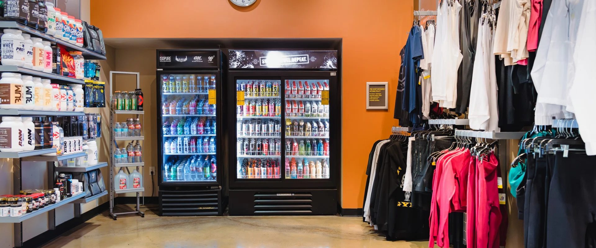 Pro Shop featuring shelves with protein powders on the left, two coolers stocked with beverages in the center, and sports apparel on racks to the right under an orange wall with a clock.