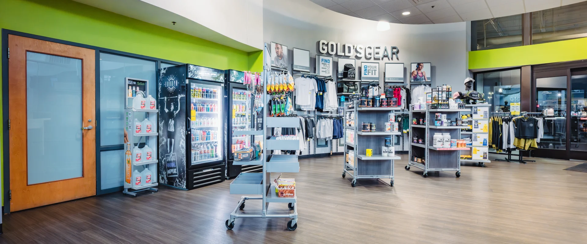 Bright Pro Shop with wooden floors, showcasing workout apparel, accessories, and supplements. Refrigerators display drinks. Modern, organized atmosphere.