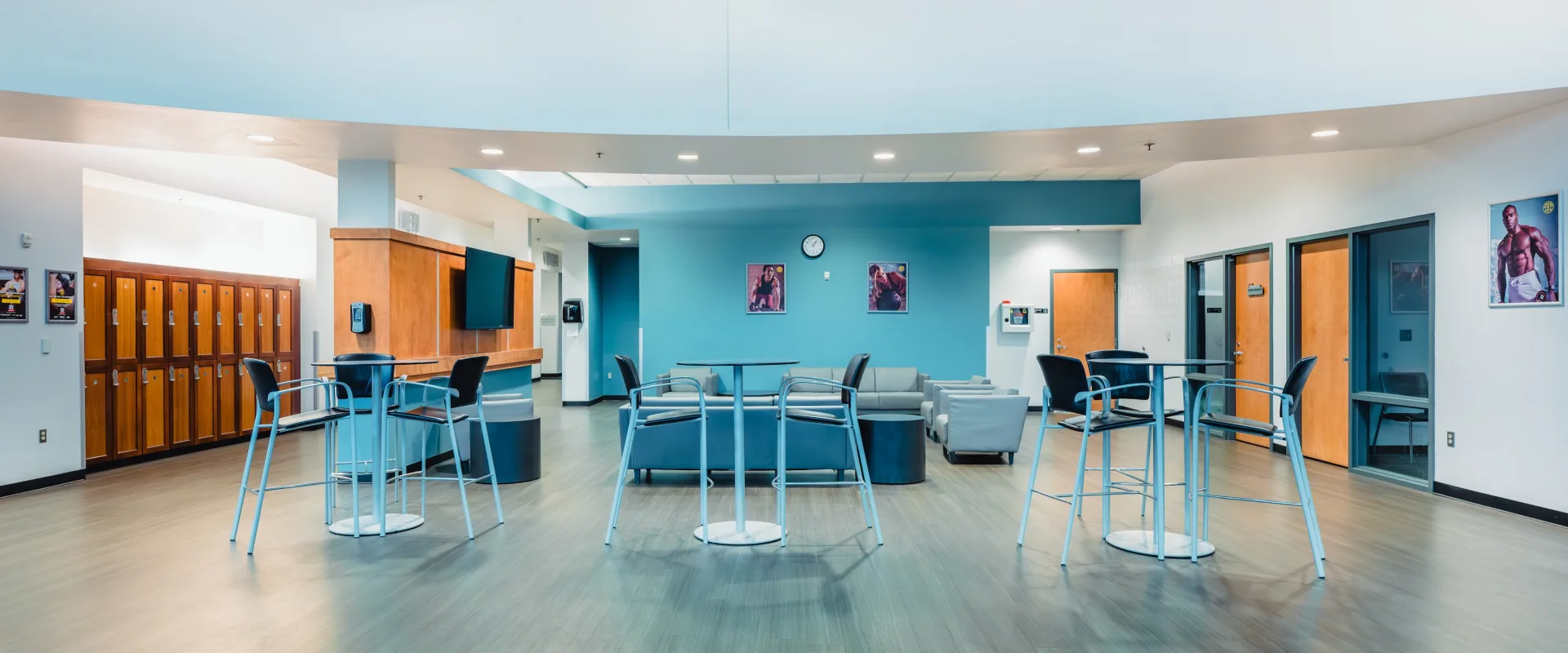 Modern lounge with blue walls, gray sofas, tall tables, and black chairs. Lockers line the left wall; doors line the right. Bright, clean ambiance.