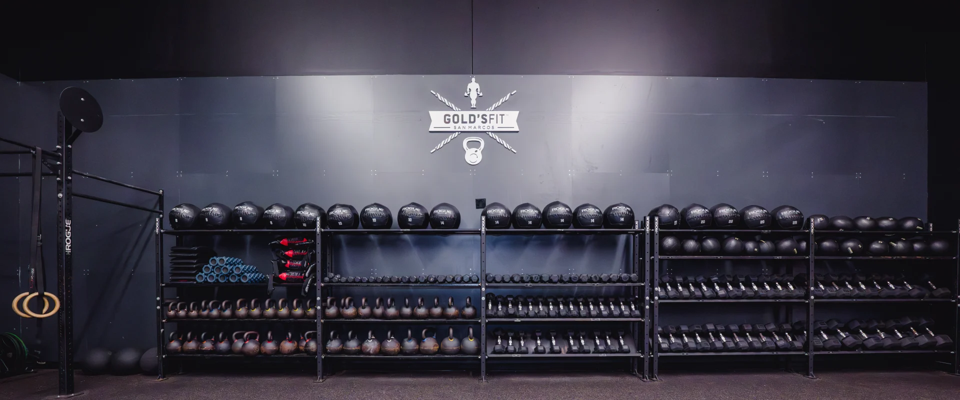 Weight shelves stocked with Rogue medicine balls, kettlebells, and dumbbells in the Gold's Fit area at Gold's Gym San Marcos, with a branded wall in the background.