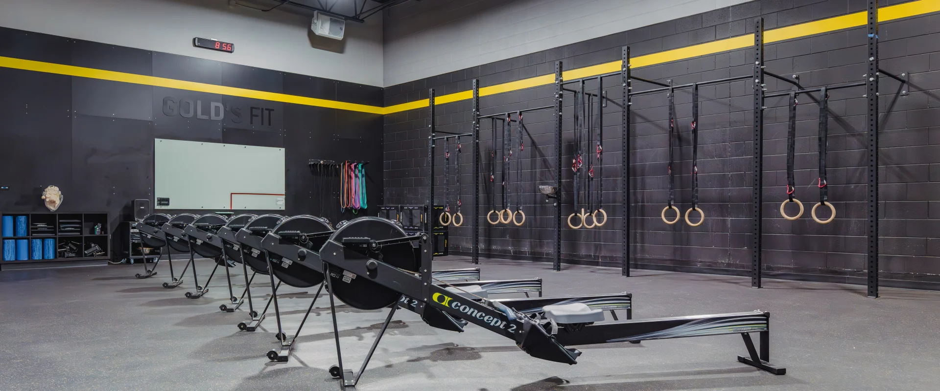 Rowing machines lined up in the Gold's Fit area, with wall-mounted gymnastic rings, resistance bands, and cubbies for equipment, against a black and yellow-accented wall.