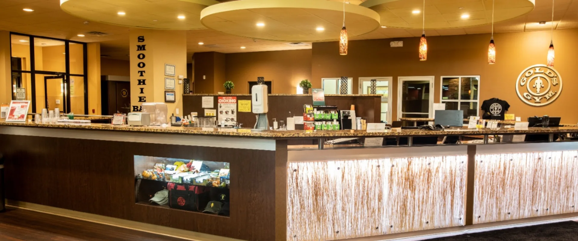 Smoothie bar at Gold’s Gym in East Northport, New York, featuring a granite countertop with a display of supplements, protein snacks, and branded merchandise. The bar includes a hand sanitizer station, blenders, and stacked cups for quick service. Pendant lights and large circular ceiling fixtures provide warm lighting, while the Gold’s Gym logo is prominently displayed on the back wall.
