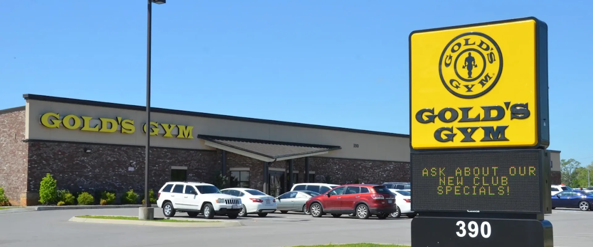 Exterior of Gold’s Gym Clarksville with yellow logo and roadside sign promoting new club specials.