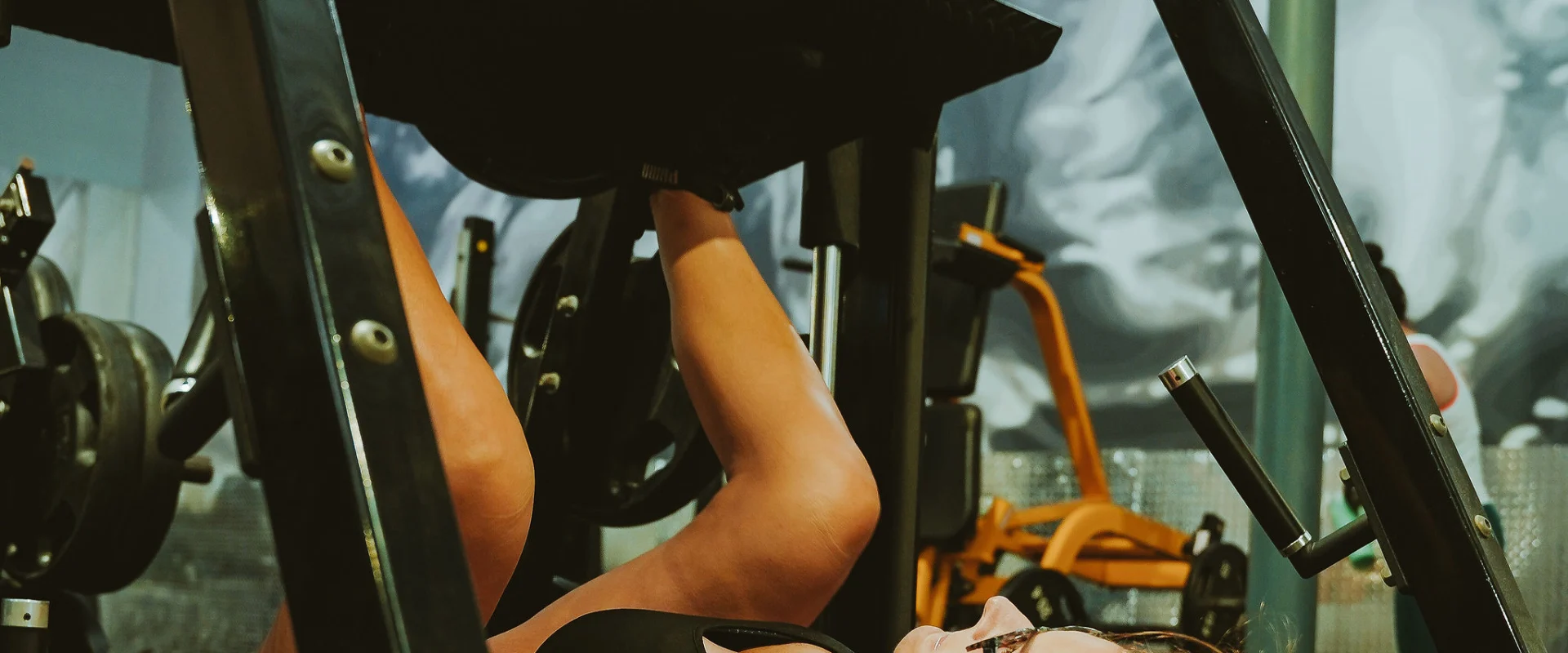 Woman performing a leg press on a plate-loaded machine at Gold's Gym Smyrna, Tennessee. The gym background includes a large mural, other exercise machines, and a lit industrial ceiling.
